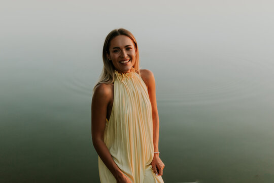 Woman stands by the water and smiles during the early morning in a light dress