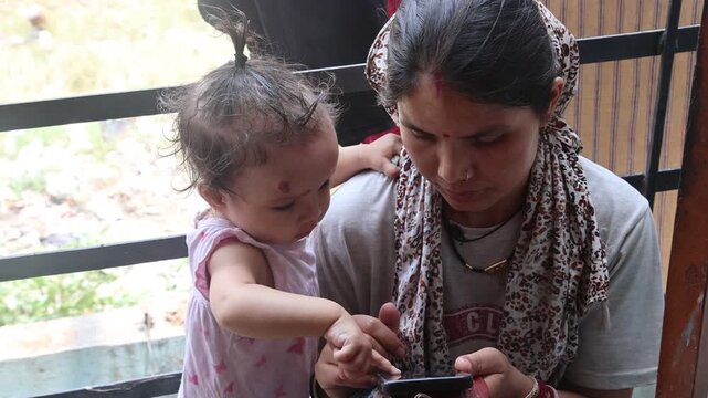 Video of an Indian mother using a smartphone while her toddler watches and scrolls with a finger. Authentic family footage capturing parenting, curiosity, learning, and everyday home life.