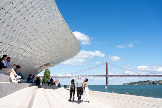 A wide shot captures visitors walking along the wide stone promenade beneath the sweeping, tiled underbelly of the MAAT (Museum of Art, Architecture and Technology) in Lisbon, Portugal.