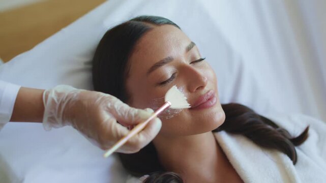 Young caucasian woman receiving creamy facial mask application with cosmetic brush, 4K
