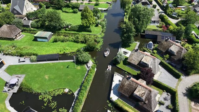 Tracking drone video of two boats navigating a narrow water canal in Wanneperveen. The camera follows the action, flanked by thatched vacation homes in the Dutch wetlands.