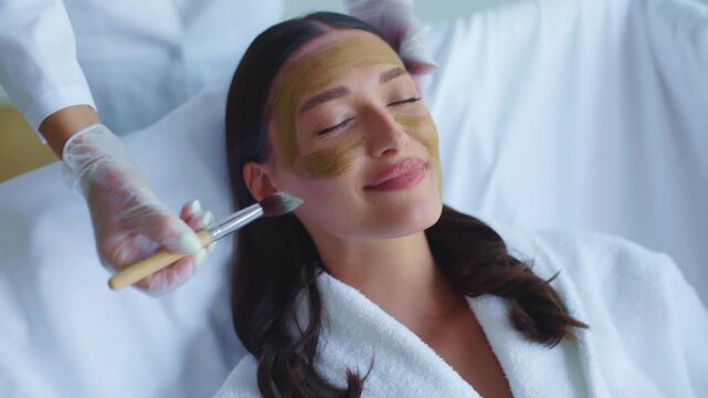 Young woman relaxing in spa while esthetician applying clay facial mask, 4K