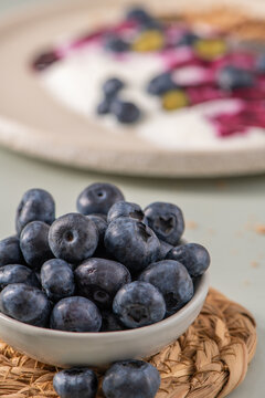 Blueberries in a small bowl with out of focus yogurt and berry topping in the background