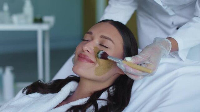 Beautician applying golden clay mask to young womans face with brush, 4K