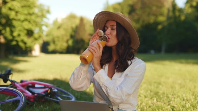 Happy lady taking break from remote work, sipping juice and gazing away with smile while sitting on blanket in green park, 4K, cinematic footage