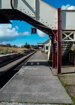 A quiet, deserted train platform at a historic railway station, characterised by its classic pedestrian footbridge and rustic setting, is waiting for passengers in a rural landscape.