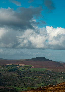 A wide, scenic view of a rolling upland landscape featuring deep-toned moorland hills, patches of bright green agricultural fields, and scattered trees, all set against a dramatic sky.