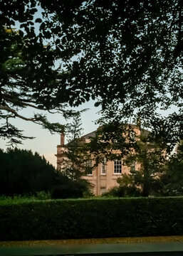 A stately, multi-story Georgian-style country house stands nestled among dense, green foliage and trees at dusk, viewed from behind a neatly trimmed hedge that frames the architectural details.