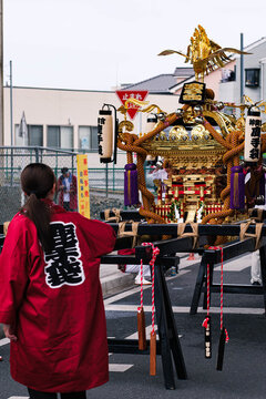 Back view of a woman in a red Happi coat standing near a golden Mikoshi portable shrine with a phoenix ornament at a Japanese festival