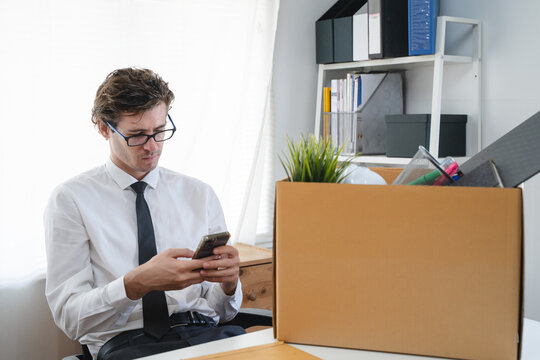 Young stressed businessman packing his belongings after being fired using smartphone in the office