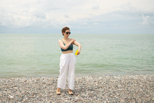 woman applying sunscreen to her shoulder by the sea, highlighting careful uv protection, healthy skincare habits and summer sun safety on a beach vacation