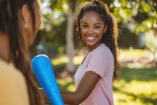 two young women in casual clothes playing with a blue plastic bat in a sunlit park with green trees and grass, playful and focused