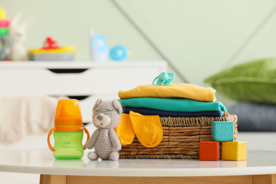 Baby clothes and accessories in a wicker basket with a sippy cup and teddy bear on a white table