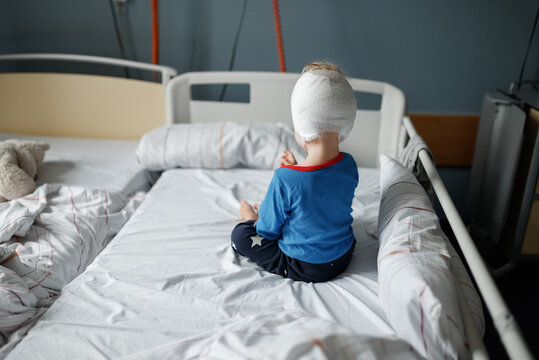 Toddler sitting in a hospital bed after cochlear implant surgery with a head bandage covering the surgical area. Rear view of a young child recovering in a pediatric hospital room after head surgery.