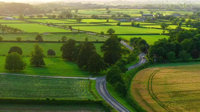 Country road countryside drive through green farmland. Aerial drone view of winding rural road with cars traveling between agricultural fields. Golden hour countryside commute across patchwork farm