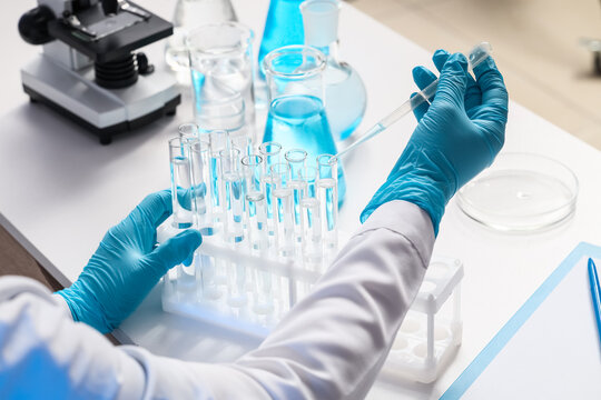 Female scientist with different flasks dropping water sample into test tube at table in laboratory, closeup