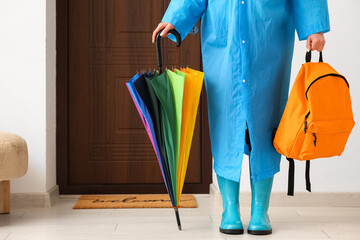 Woman in raincoat and gumboots holding umbrella with backpack near door at home, closeup © Pixel-Shot