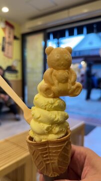 Hand holding a yellow soft serve ice cream in a waffle cone topped with a cute teddy bear monaka