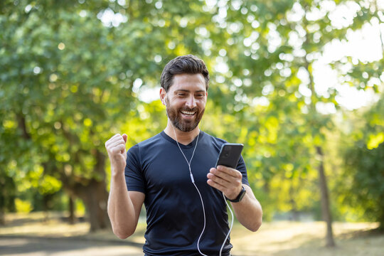 Happy young male athlete standing in headphones on the street, looking at his mobile phone and rejoicing in victory, showing a gesture with his hand