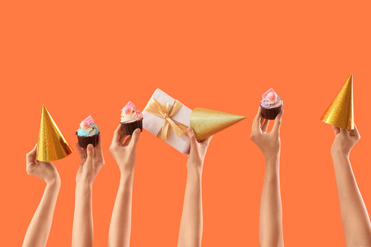 Many female hands with birthday cupcakes, gift box and party hats on orange background