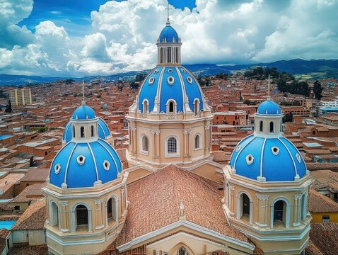 Aerial view of a majestic church with three bright blue domes overlooking terracotta rooftops and distant mountains beneath a dramatic cloud-filled sky, evoking serenity