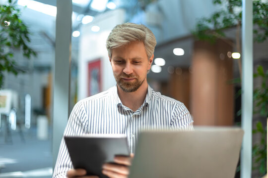 Smiling busy young employee, young entrepreneur using tablet computer, happy businessman executive looking at tab device analyzing finance trading market data working sitting at office desk.
