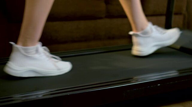 Unrecognizable person walking on treadmill belt and gradually stopping during indoor cardio session. White sneakers stepping along moving track slowing pace in cozy apartment workout space. Home