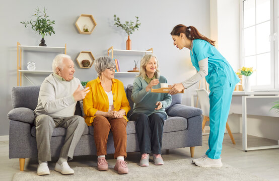 Friendly young nurse serving tea and snacks to group of cheerful seniors sitting on cozy sofa in retirement home. Joyful elderly pensioners relaxing together with care and companionship of caregiver.