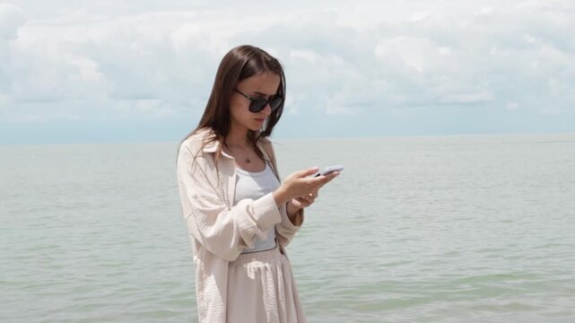 young woman in sunglasses uses a smartphone beside the sea, combining mobile technology, travel planning, online communication and a relaxed beach vacation by the water