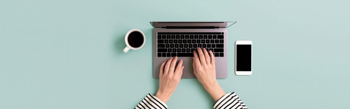 Laptop and computer workspace with hands typing near coffee and smartphone on desk