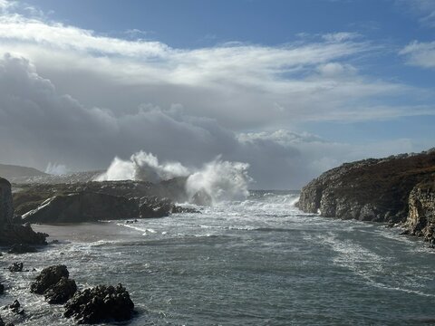 Powerful Atlantic waves crash against a rocky coastline beneath a dramatic storm front as blue sky breaks through the clouds. Explosive surf and sea spray reveal the raw force of the ocean.