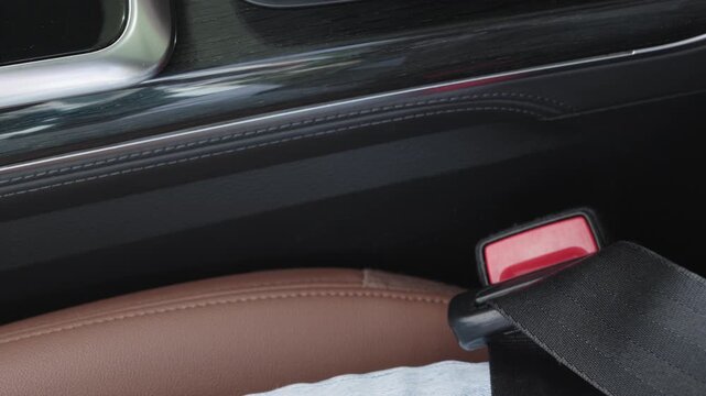 Close-up of passenger hand inserting seat belt tongue into red buckle receiver next to stitched brown leather seat inside modern car interior.