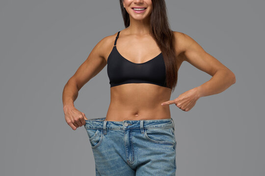 Smiling athletic woman in black sports bra proudly showing loose jeans after fitness transformation