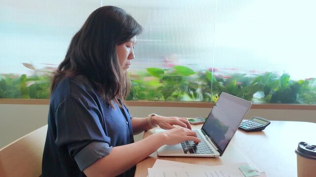 A business professional works on a laptop at a tidy desk while reviewing printed documents in a comfortable office.