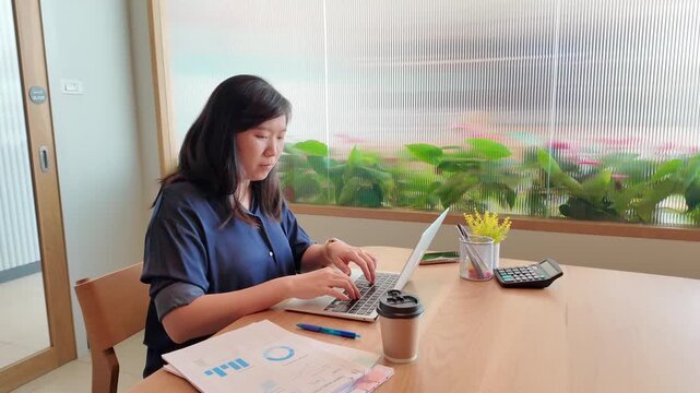 A business professional works on a laptop at a tidy desk while reviewing printed documents in a comfortable office.