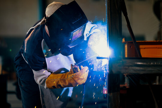 Welder wears a helmet and gloves. Bright sparks fly from the welding torch. He focuses intensely on the metal work. Industrial equipment surrounds him. This captures skilled craftsmanship