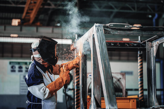 Industrial worker welds metal framework with sparks flying. Worker wears protective gear, focused expression. Factory setting shows skilled labor in manufacturing environment. Ideal for industrial