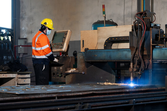 Worker wears a yellow hard hat and orange safety vest. He stands near machinery with blue sparks flying. Metal sheets cover the factory floor. The worker concentrates on equipment operation