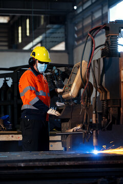 Worker in industrial attire handles welding equipment. He wears a yellow hard hat and an orange safety vest. Blue sparks fly as he welds metal. He uses a face mask and focuses on the task