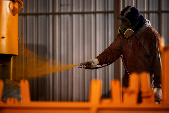 Worker sprays orange paint in a factory setting. Wearing a helmet and gas mask, the person concentrates on the task. Metal structures and corrugated walls form the background