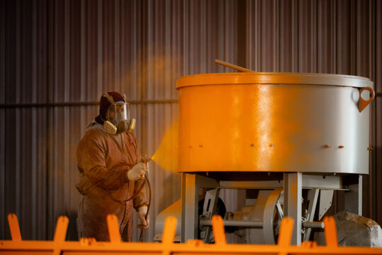 Worker in full hazmat suit sprays industrial container. Dust clouds form around the large metallic vessel. The worker wears a helmet and protective eyewear. Concentrated focus guides the spray nozzle