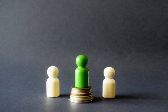 Studio still life of group dynamics and hierarchy featuring a central green peg doll standing on a coin stack between two lower-positioned plain wood figures against a dark grey background.
