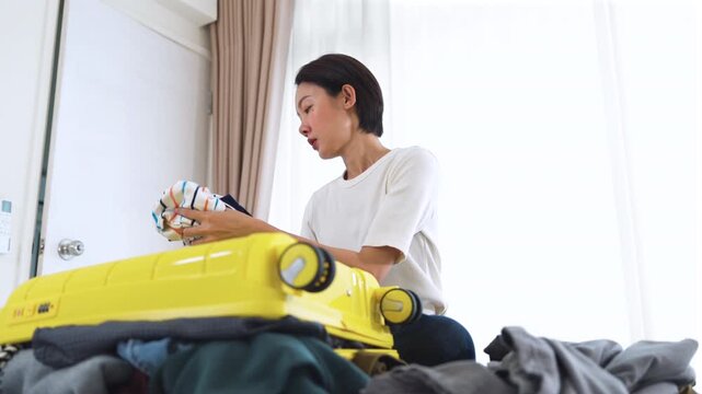 Exhausted Asian female tourist having a headache behind a messy bursting luggage. Concept of heavy baggage, overpacking issues and vacation planning stress at home.