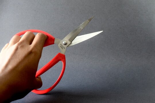 A close-up shot of a hand grasping a pair of red plastic-handled stainless steel scissors against a solid dark gray surface, angled for copy space on the right.