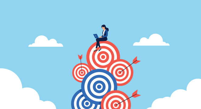 Businesswoman sitting on a tall stack of red and blue bullseye targets while working on a laptop to represent goal achievement.