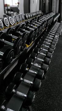 Diagonal rack of chrome handled rubber dumbbells in clean commercial gym interior, showing orderly rows of round weights, mirrored reflections, cool lighting, and strong perspective for fitness