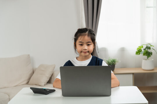 Young girl sitting at modern white desk using laptop computer with calculator nearby in bright home interior focusing on screen with neutral expression during daytime