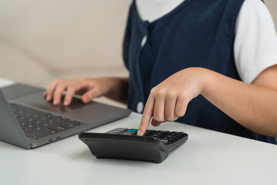 Child using calculator and laptop on white desk for learning numerical skills with focused hand pressing button in education and technology setting