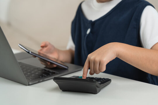 Child using calculator and holding smartphone while sitting in front of laptop working on math homework or financial calculation at white desk indoors