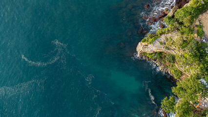 Drone top-down view of a striking rip current pattern in deep turquoise ocean water, framed by a rugged tropical cliff with waves breaking against the rocks. © Fredy